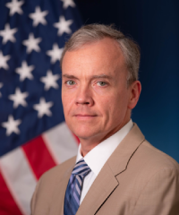 A headshot of Dr. Timothy Grayson in a tan suit with a navy tie infront of an American flag